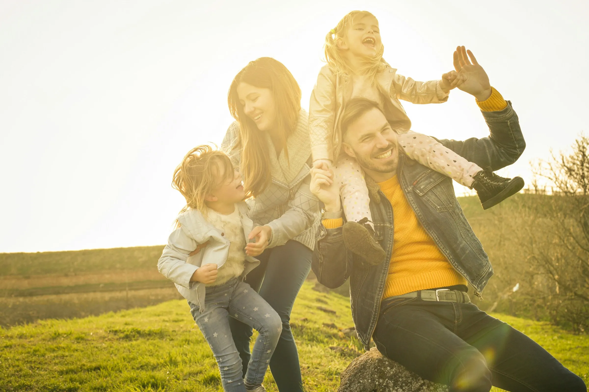 A family playing together in a sunlit field, one child on a parent's shoulders, everyone laughing